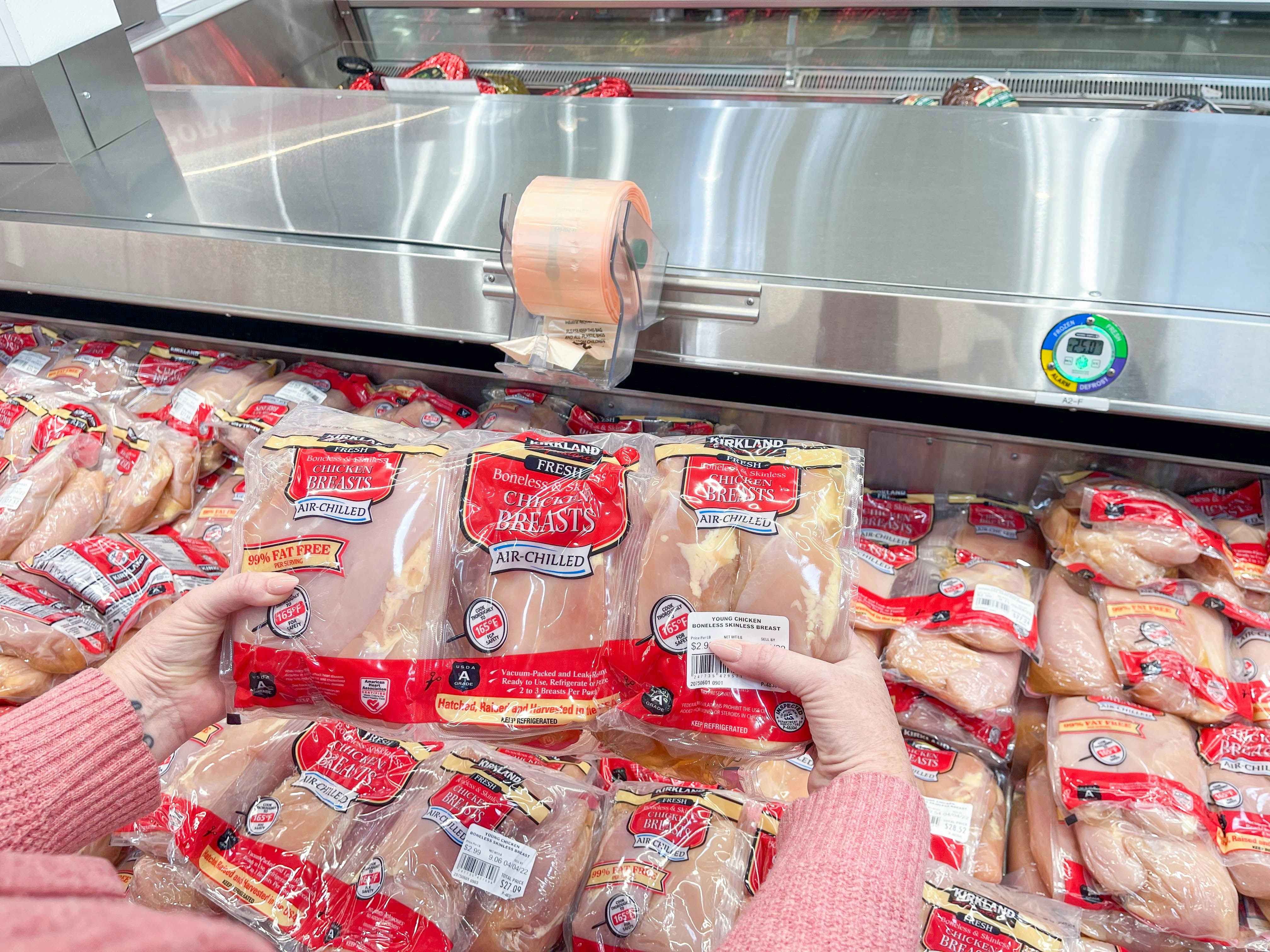 A person's hands holding up a three-pack of Kirkland raw chicken breasts above a cooler full of the same product inside Costco.