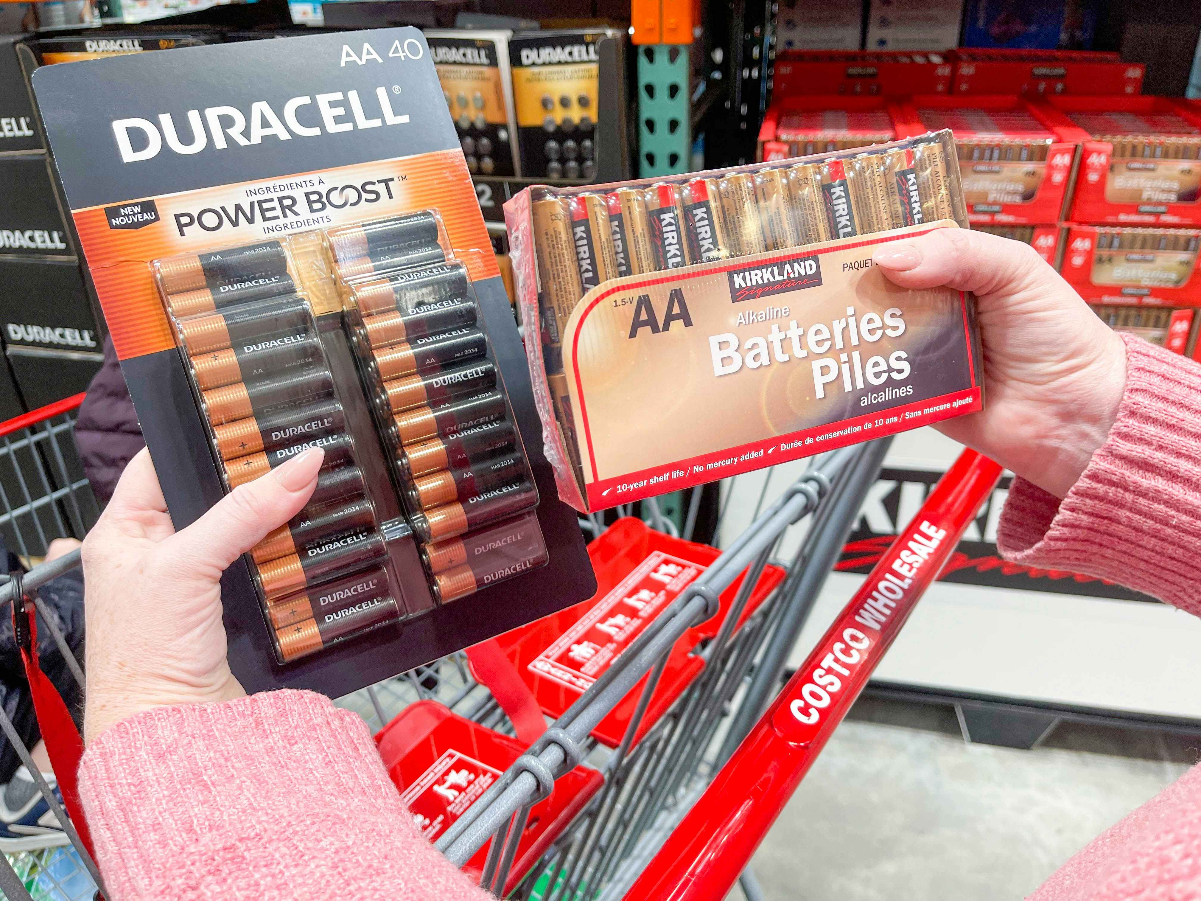 A person's hands holding up a 40-pack of Duracell AA batteries next to a a pack of Kirkland brand AA batteries above a Costco shopping cart in an aisle at Costco.