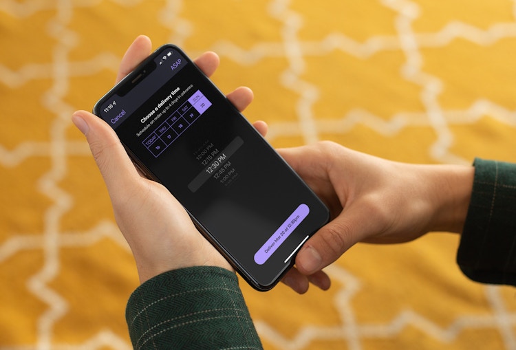 - A person holding an iPhone that is displaying the Grubhub mobile app's scheduling page. The user is setting up a delivery for a future time and date.