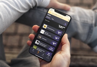 A person sitting and holding their iPhone with the Grubhub mobile app open showing the list of Grubhub Perks for different restaurants including Buffalo wild Wings, Mai Thai Restaurant & Bar, and Wild Burger. The user's Perks cash value shows to be $107.00.