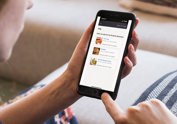- A person holding their iPhone with the Grubhub mobile app open to the Help section. The user's orders are displayed below the instructions which read, 