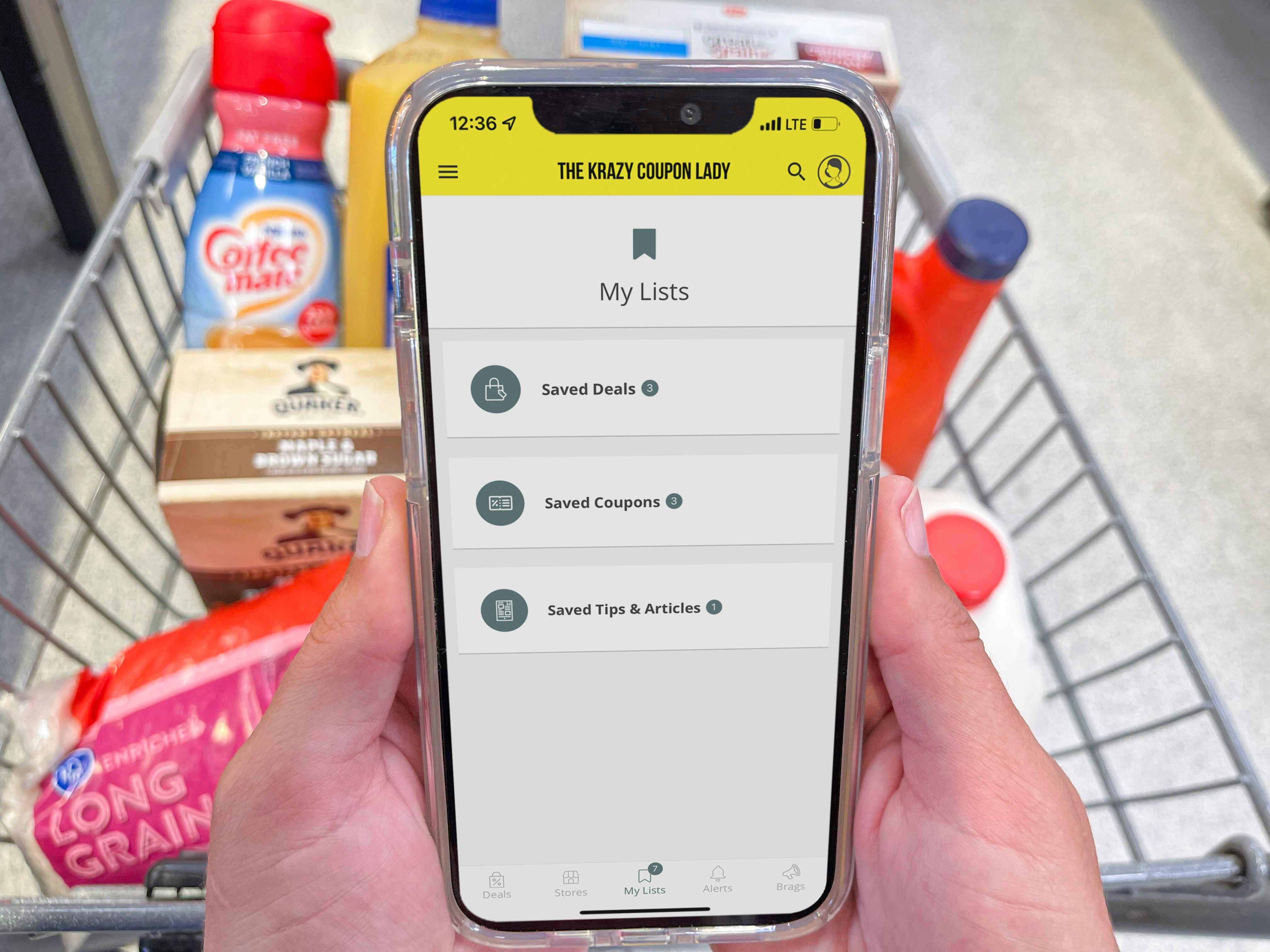 A person's hands holding an iPhone displaying the Krazy Coupon Lady mobile app's My Lists page in front of a cart filled with groceries.