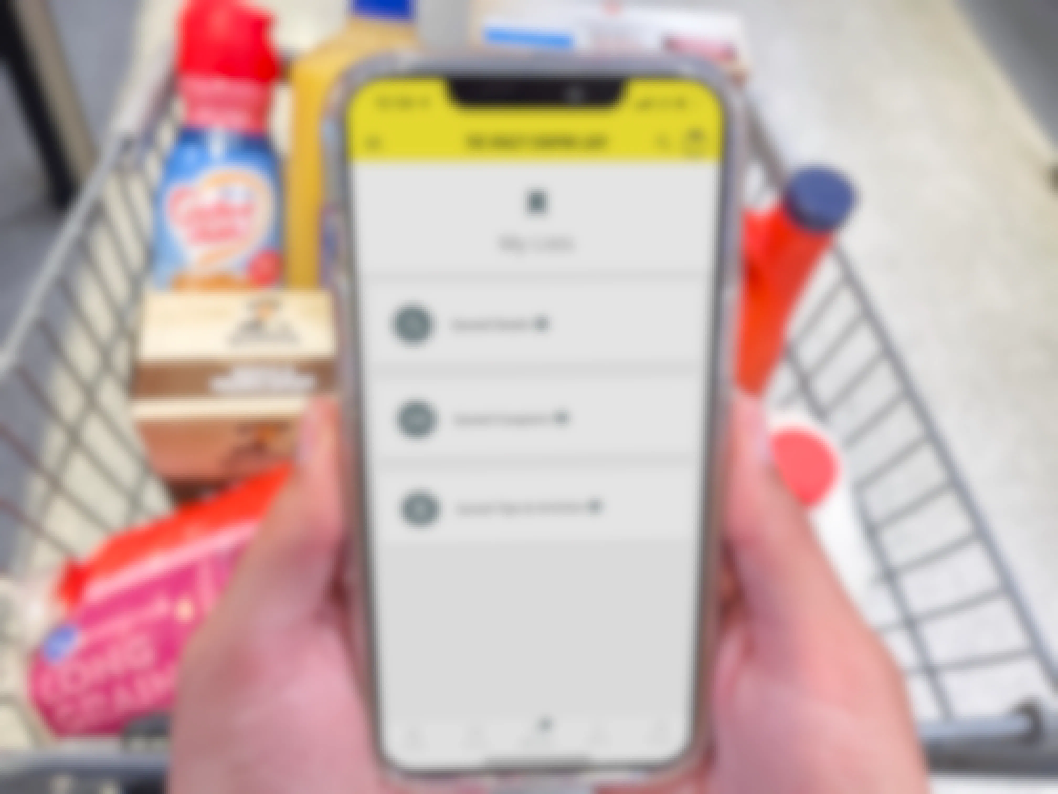 A person's hands holding an iPhone displaying the Krazy Coupon Lady mobile app's My Lists page in front of a cart filled with groceries.