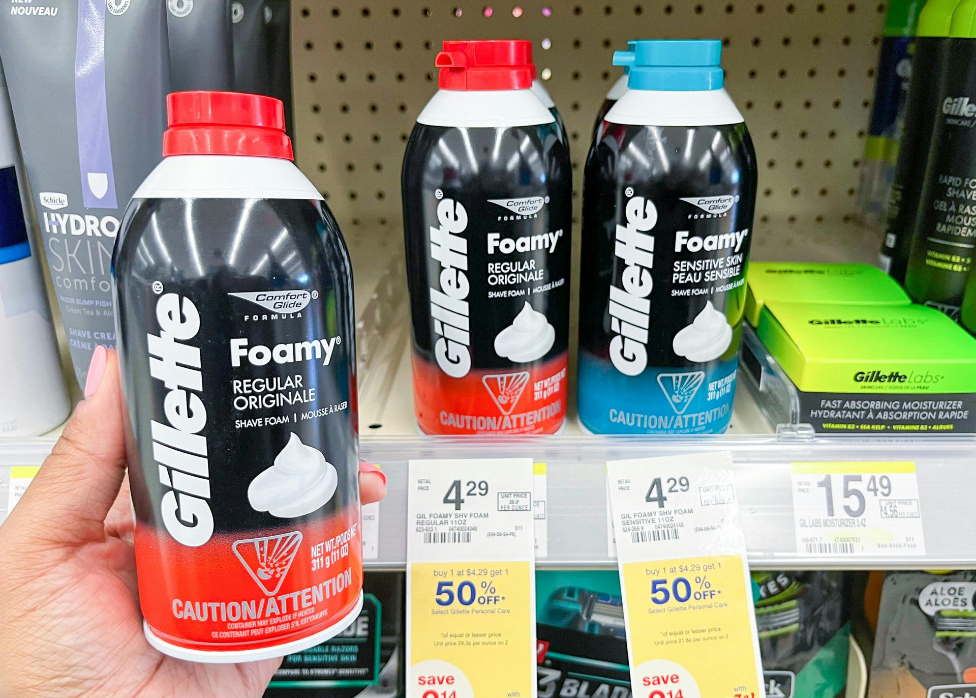 A person's hand holding up a bottle of Gillette Foamy shaving cream in front of a shelf with more Gillette shaving cream at Walgreens.