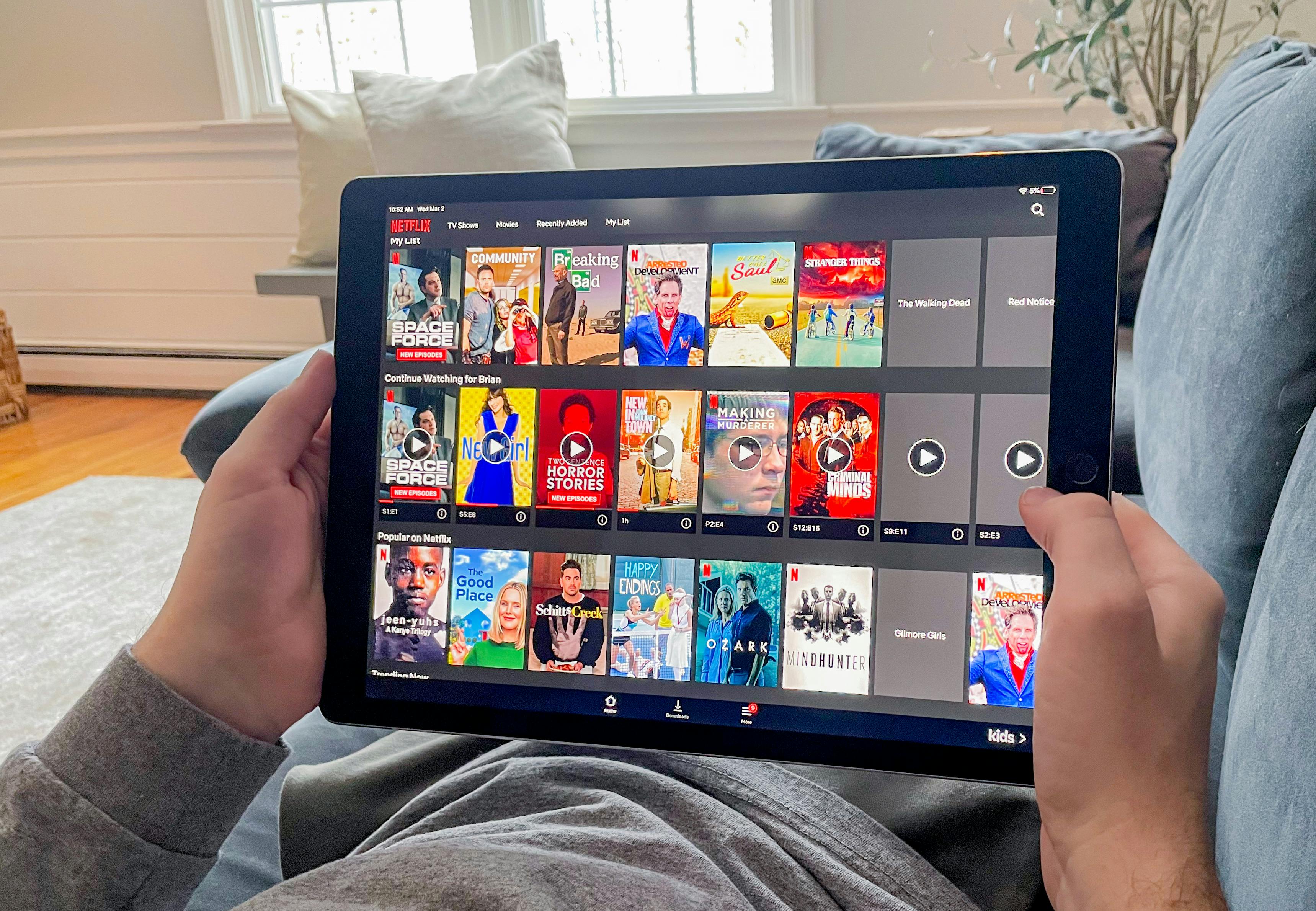 man laying on couch looking at a tablet displaying Netflix movie and show titles