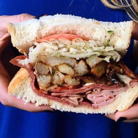 A person holding a sandwich that has sliced meats, tomato, french fries, and lettuce on it.