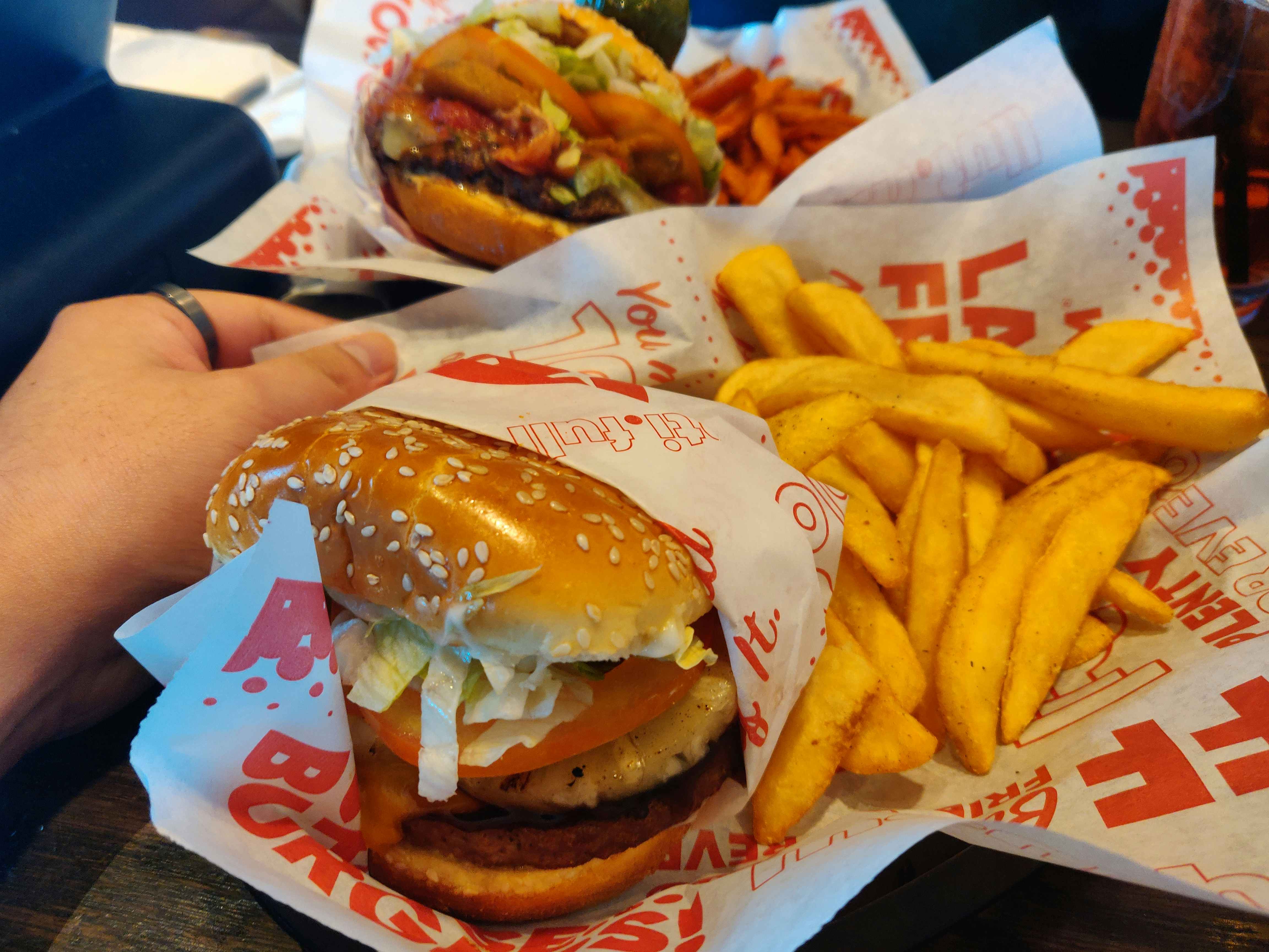 A persons hand holding a basket from Red Robin with a burger and fries with another similar basket on the table in the background.