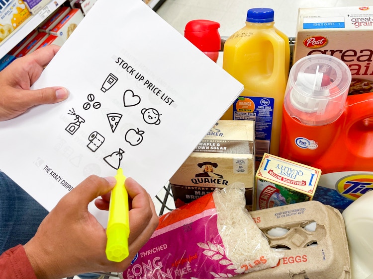 A person's hands holding a highlighter and a print-out of KCL's Stock Up Price List in front of a cart full of groceries.