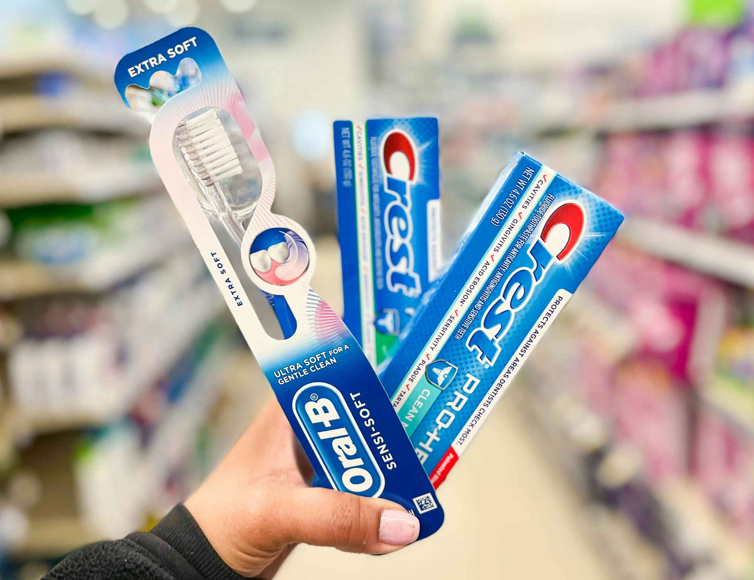 hand holding two tubes of Crest toothpaste and an Oral-B manual toothbrush in aisle