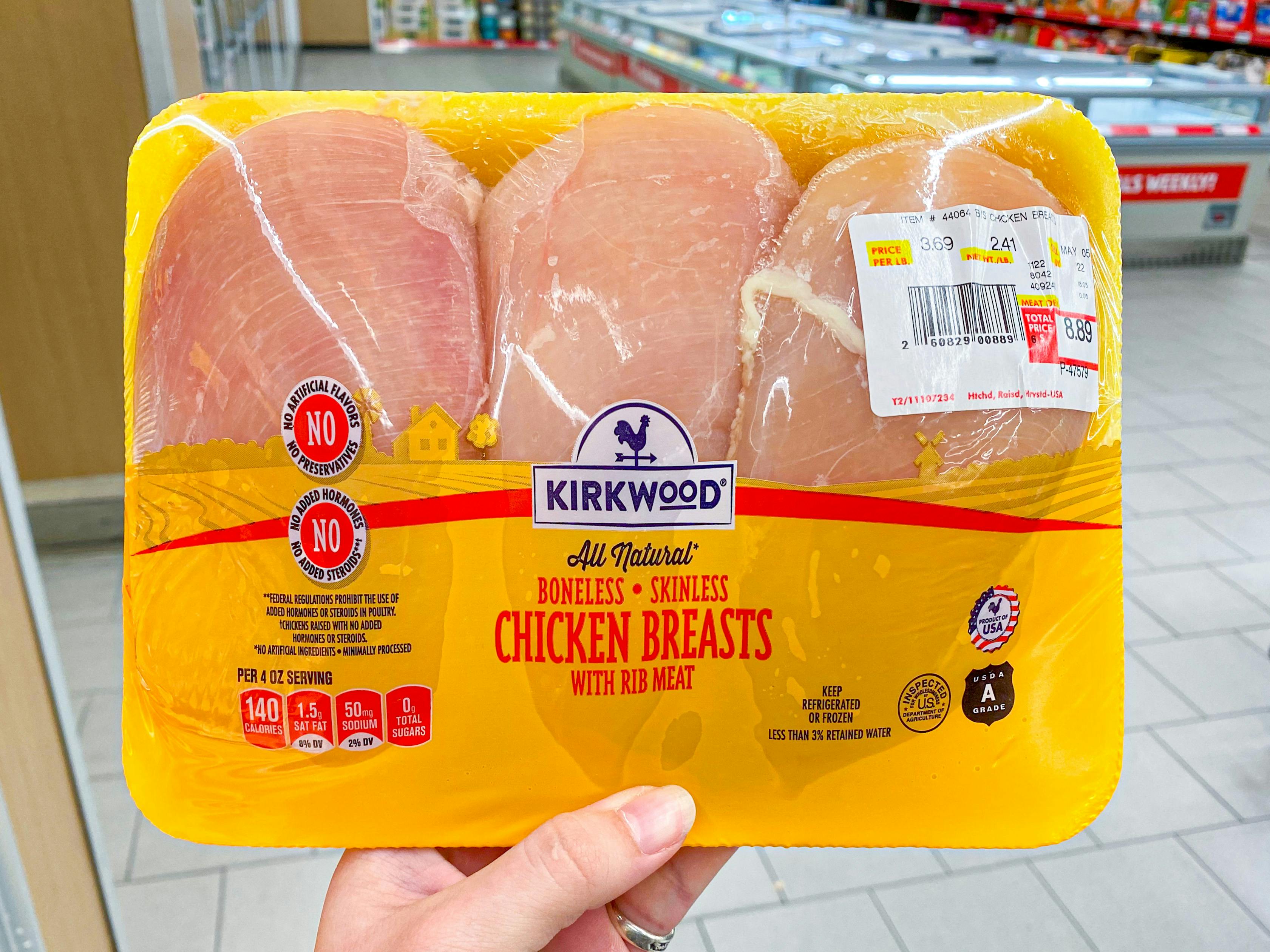 A person's hand holding a package of Kirkwood skinless, boneless chicken breasts in the refrigerated section at Aldi.
