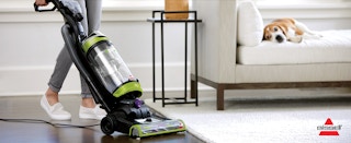 A person pushing a Bissell vacuum cleaner across a floor next to a dog laying on a couch.