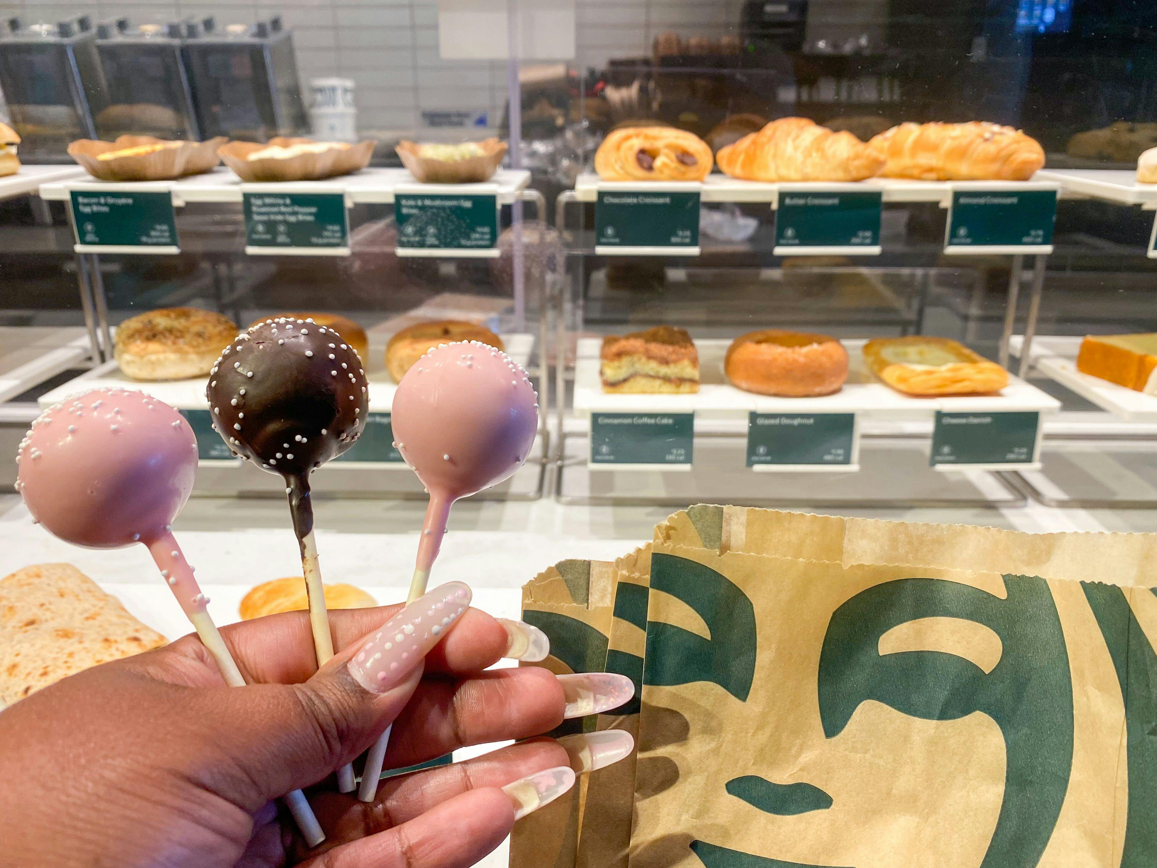 A person's hand holding up three Starbucks cake pops in front of the enclosed food display at a Starbucks counter.