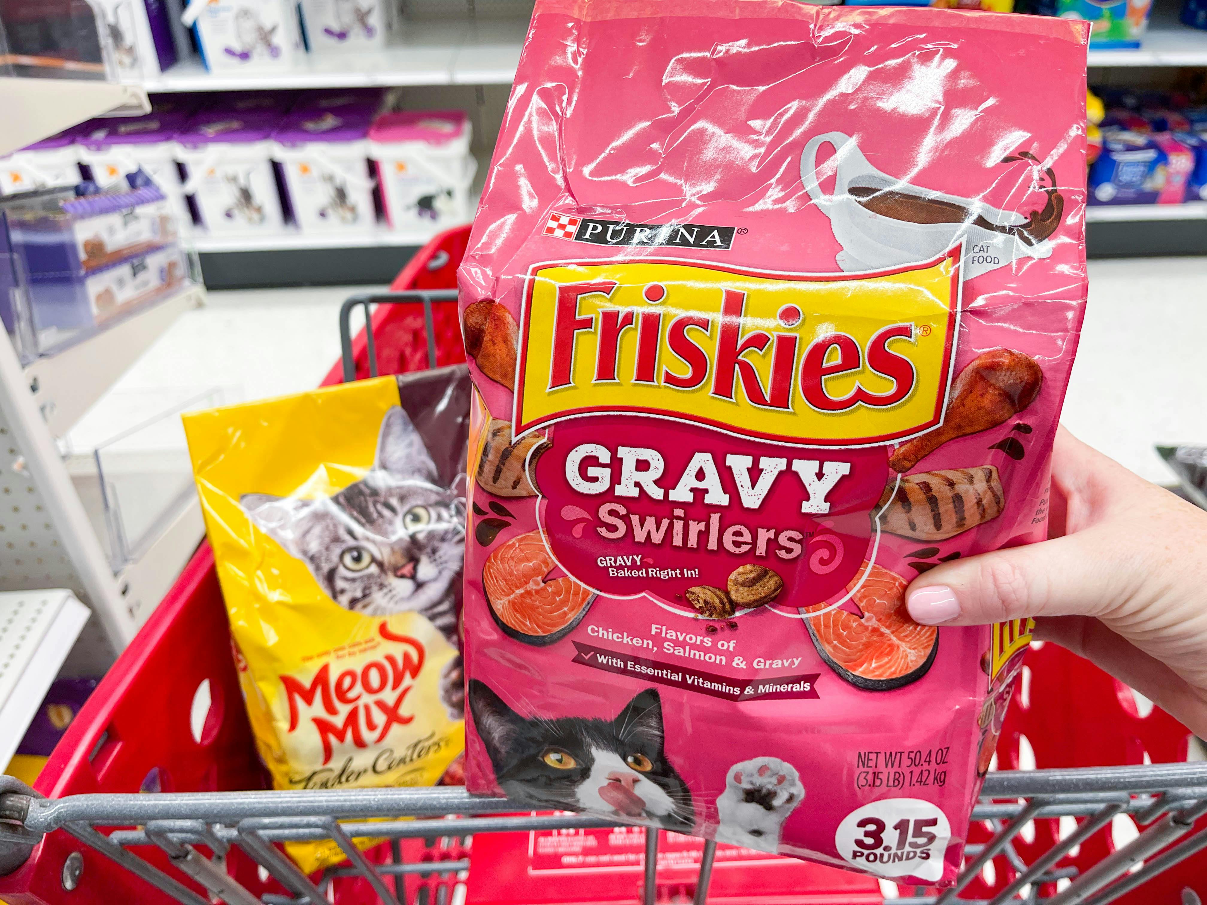 a bag of meow mix in cart in the background with a bag of friskies cat food being held on handle of cart