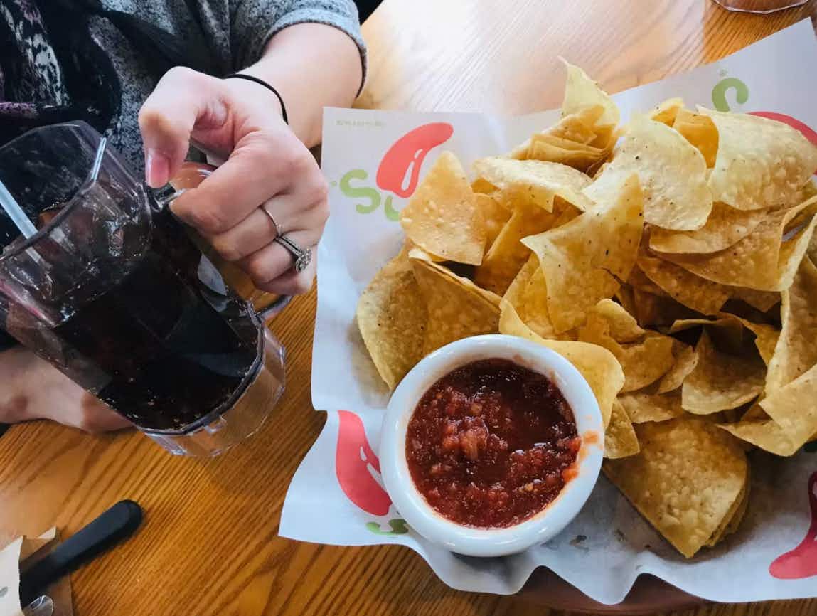 A person holding a drink glass next to a basket of Chili's chips and salsa.