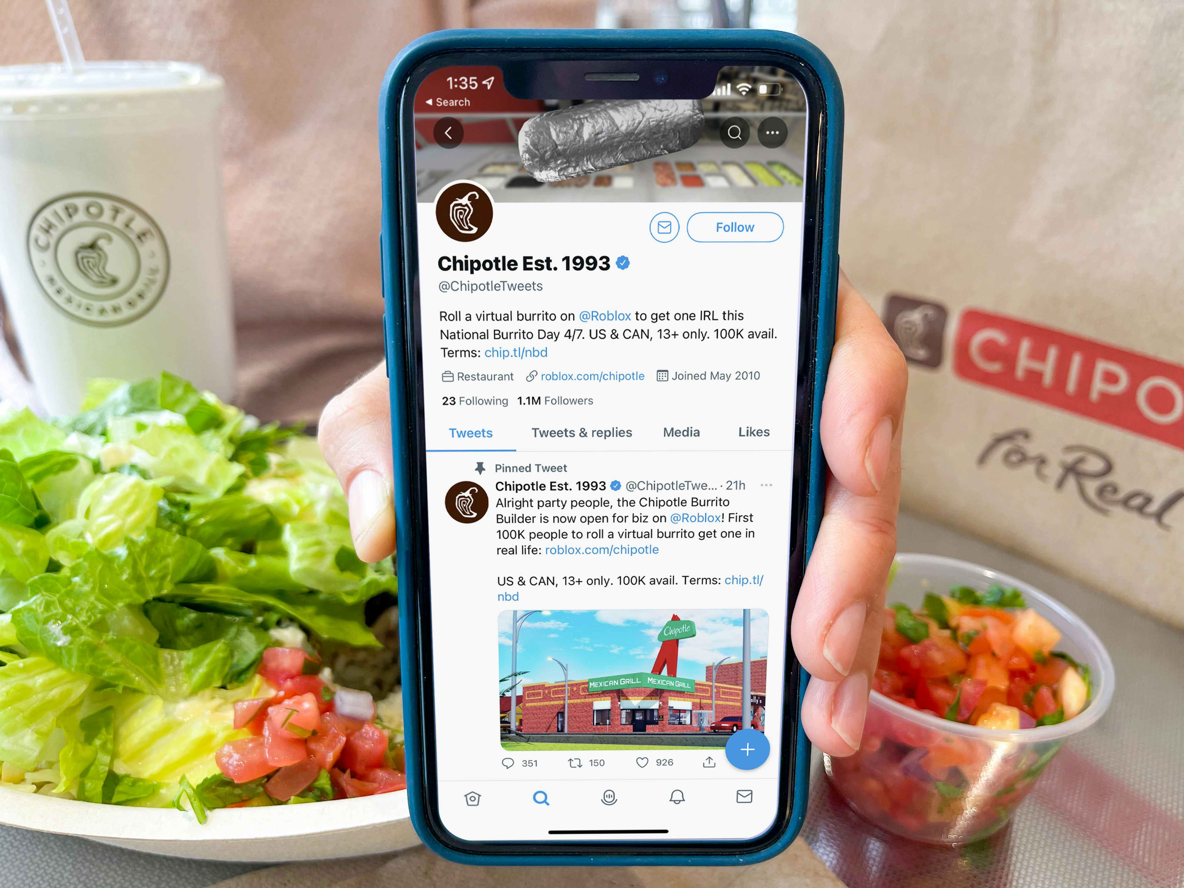 A person's hand holding up a phone displaying the Chipotle Twitter page in front of Chipotle food and takeout bag on a table.