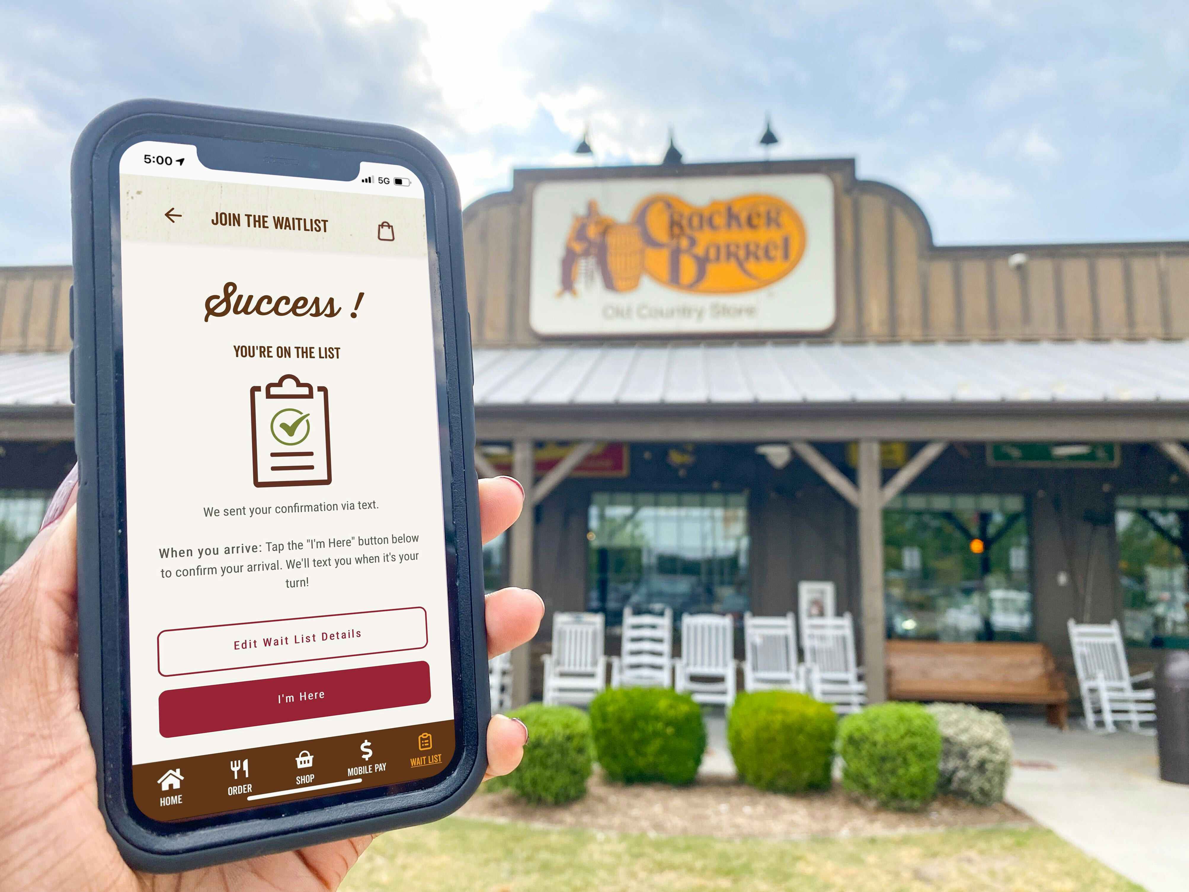 A person holding a cell phone in front of a Cracker Barrel restaurant. The waitlist section of the Cracker Barrel app is visible on the cell phone screen.