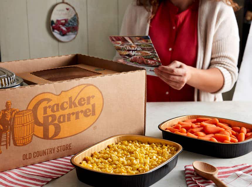 A woman looking at a pamphlet next to a Cracker Barrel catering box and pans of food on a table.