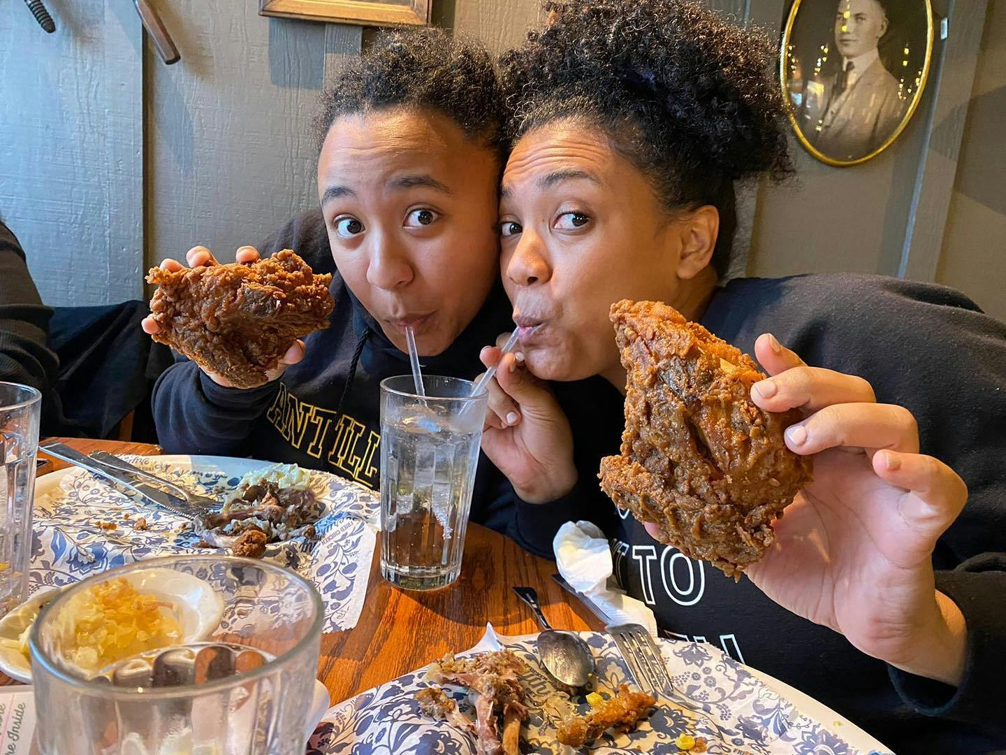 Two women sitting at a table in Cracker Barrel, drinking from the same cup with two different straws, holding up fried chicken.