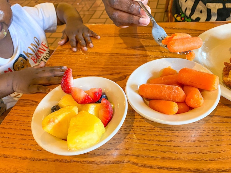 Two small bowls of mixed fruit and baby carrots sitting on a table in front of a baby with an adult picking up some carrots with a fork.