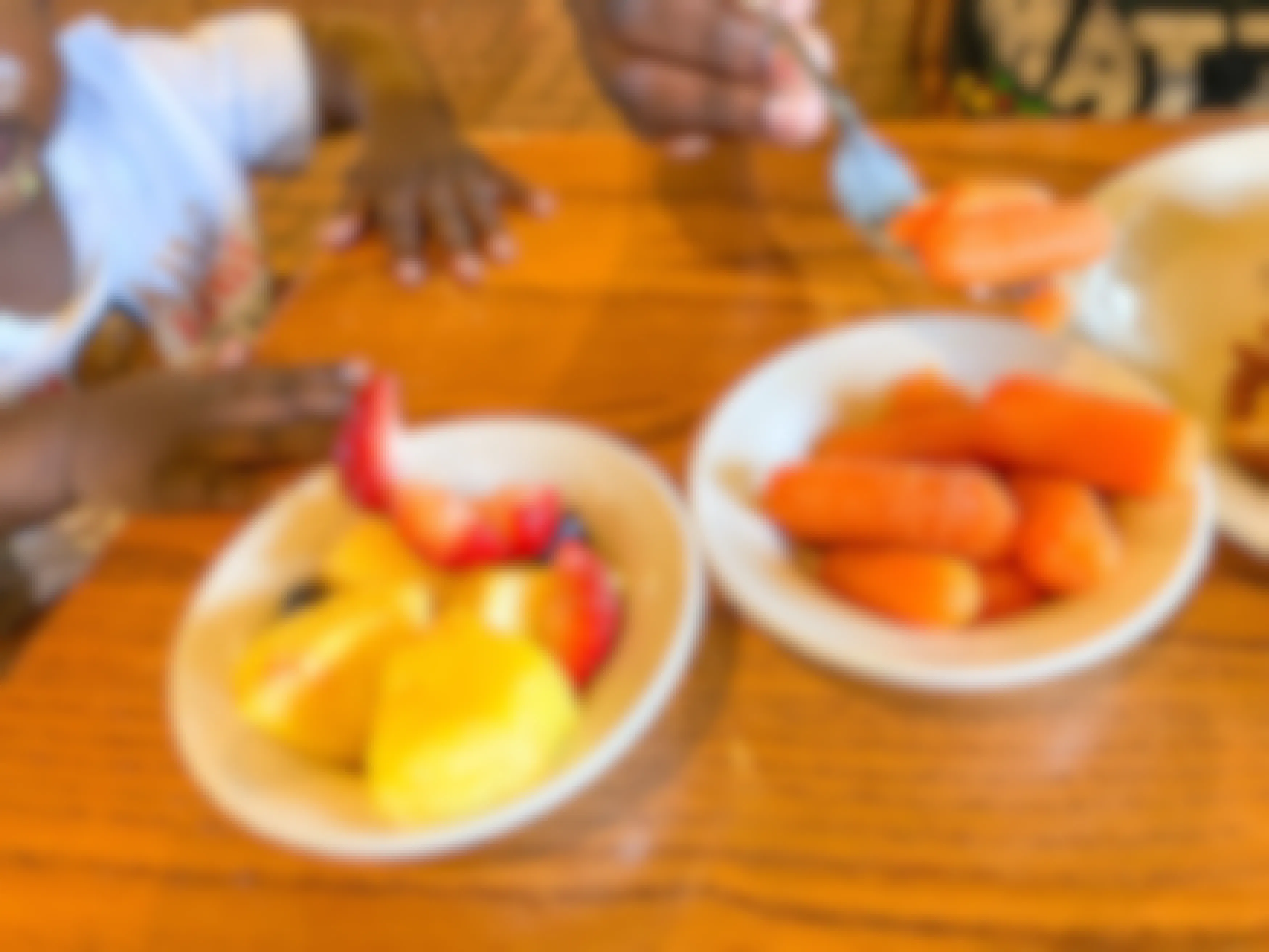 Two small bowls of mixed fruit and baby carrots sitting on a table in front of a baby with an adult picking up some carrots with a fork.