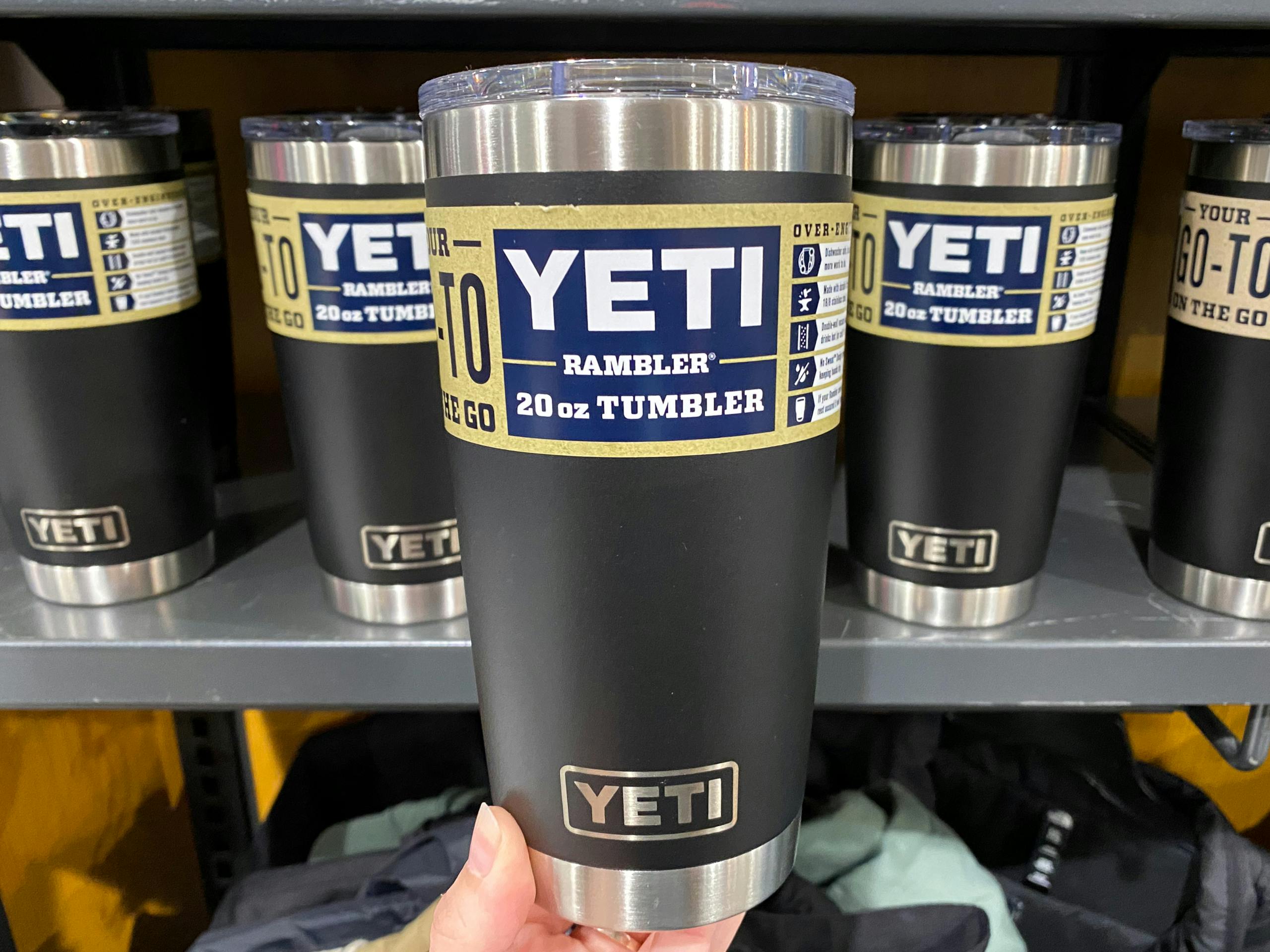 A person's hand holding up a black Yeti Rambler 20oz tumbler in front of a shelf of more of the same tumbler at Dick's Sporting Goods.