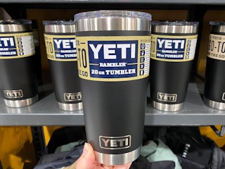 A person's hand holding up a black Yeti Rambler 20oz tumbler in front of a shelf of more of the same tumbler at Dick's Sporting Goods.