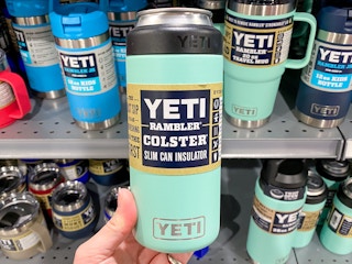 A person's hand holding up a teal Yeti Rambler Colster Slim Can Insulator in front of a shelf of more Yeti products at Dick's Sporting Goods.