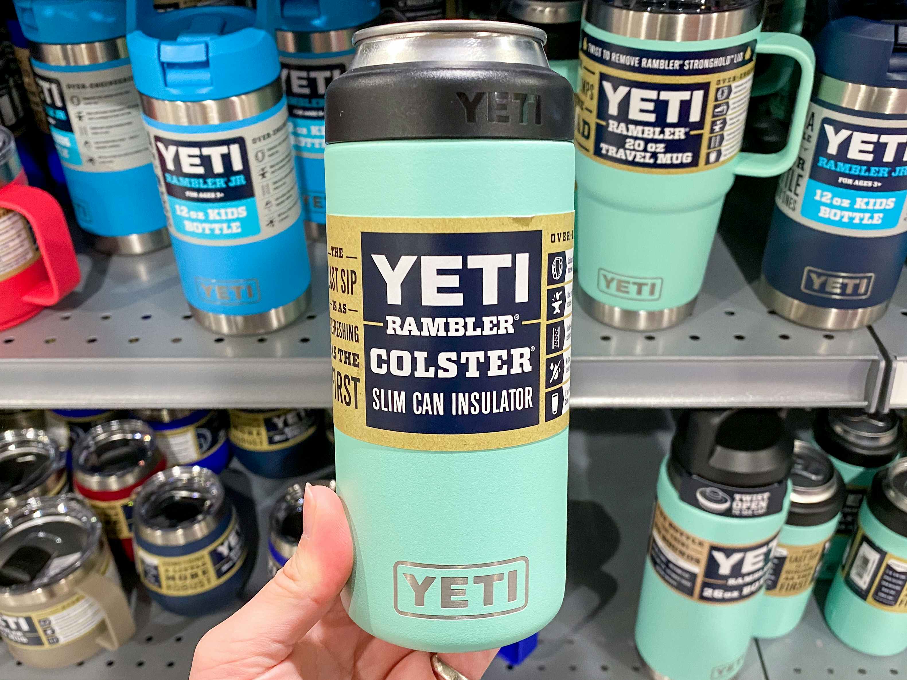 A person's hand holding up a teal Yeti Rambler Colster Slim Can Insulator in front of a shelf of more Yeti products at Dick's Sporting Goods.