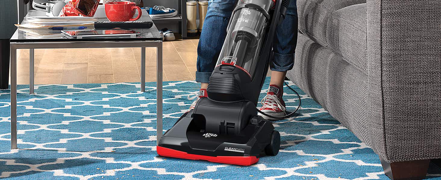 A person pushing a Dirt Devil vacuum across a patterned rug near a couch and coffee table.