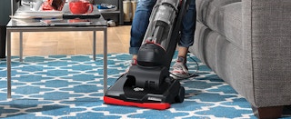 A person pushing a Dirt Devil vacuum across a patterned rug near a couch and coffee table.