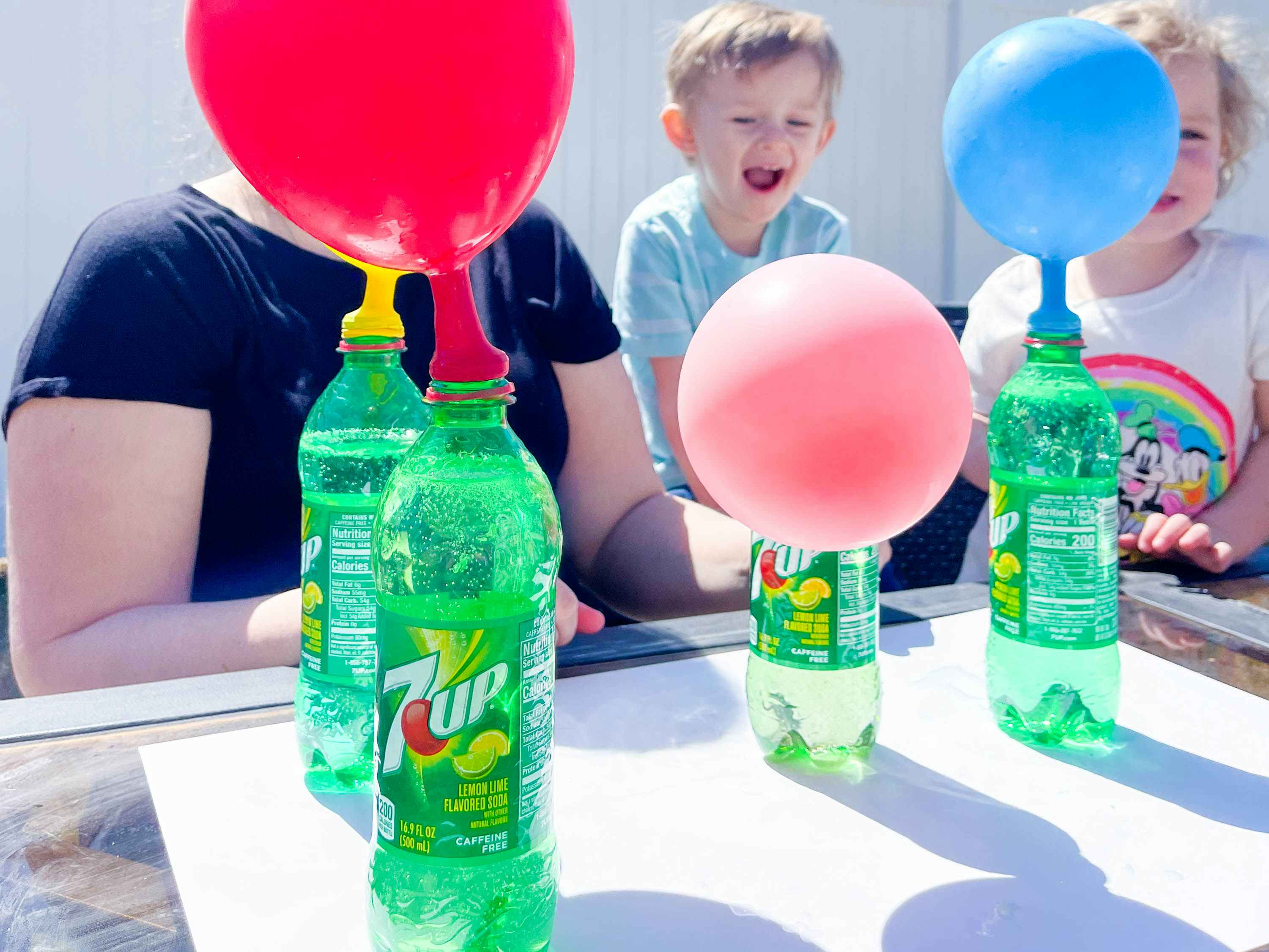 a woman and kids outside at a table with soda bottles with balloons on top to do an experiment 