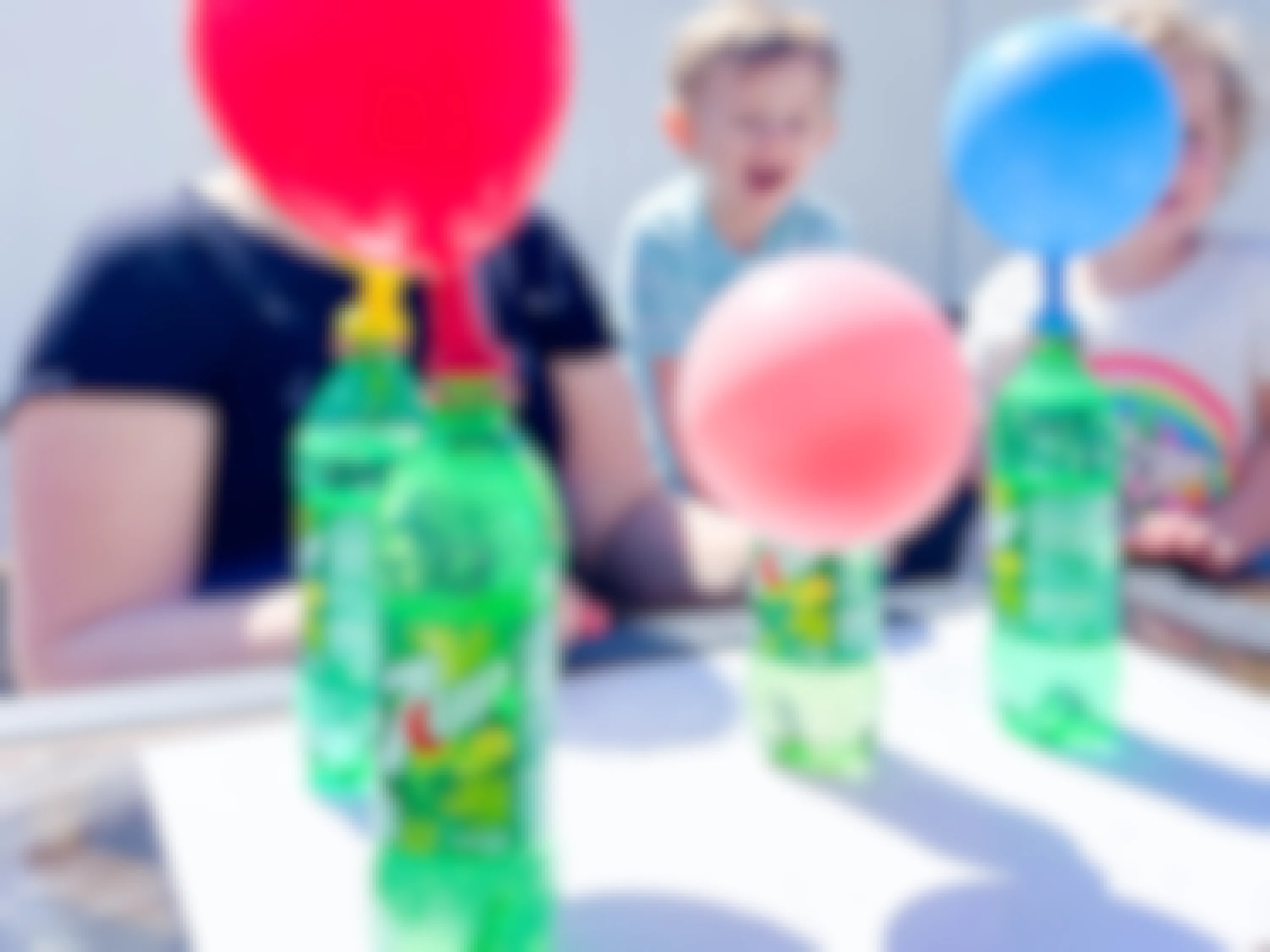 a woman and kids outside at a table with soda bottles with balloons on top to do an experiment 