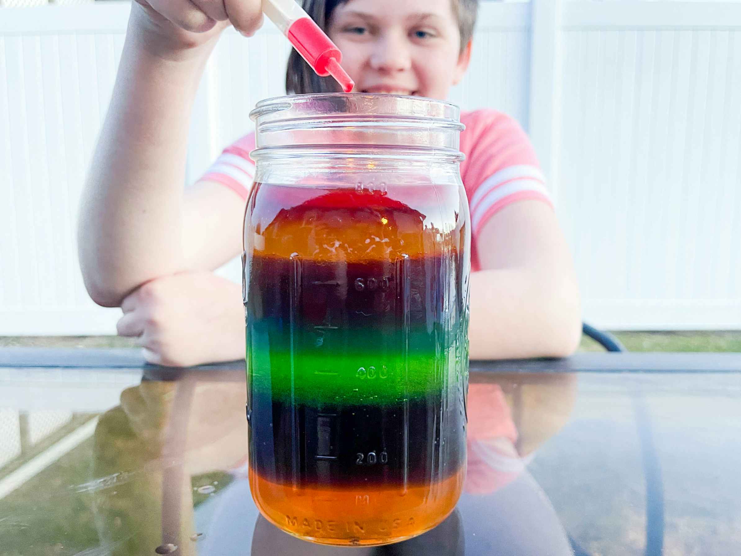 a kid holding a syringe with liquid over a mason jar filled with mulitple liquids to create a rainbow look