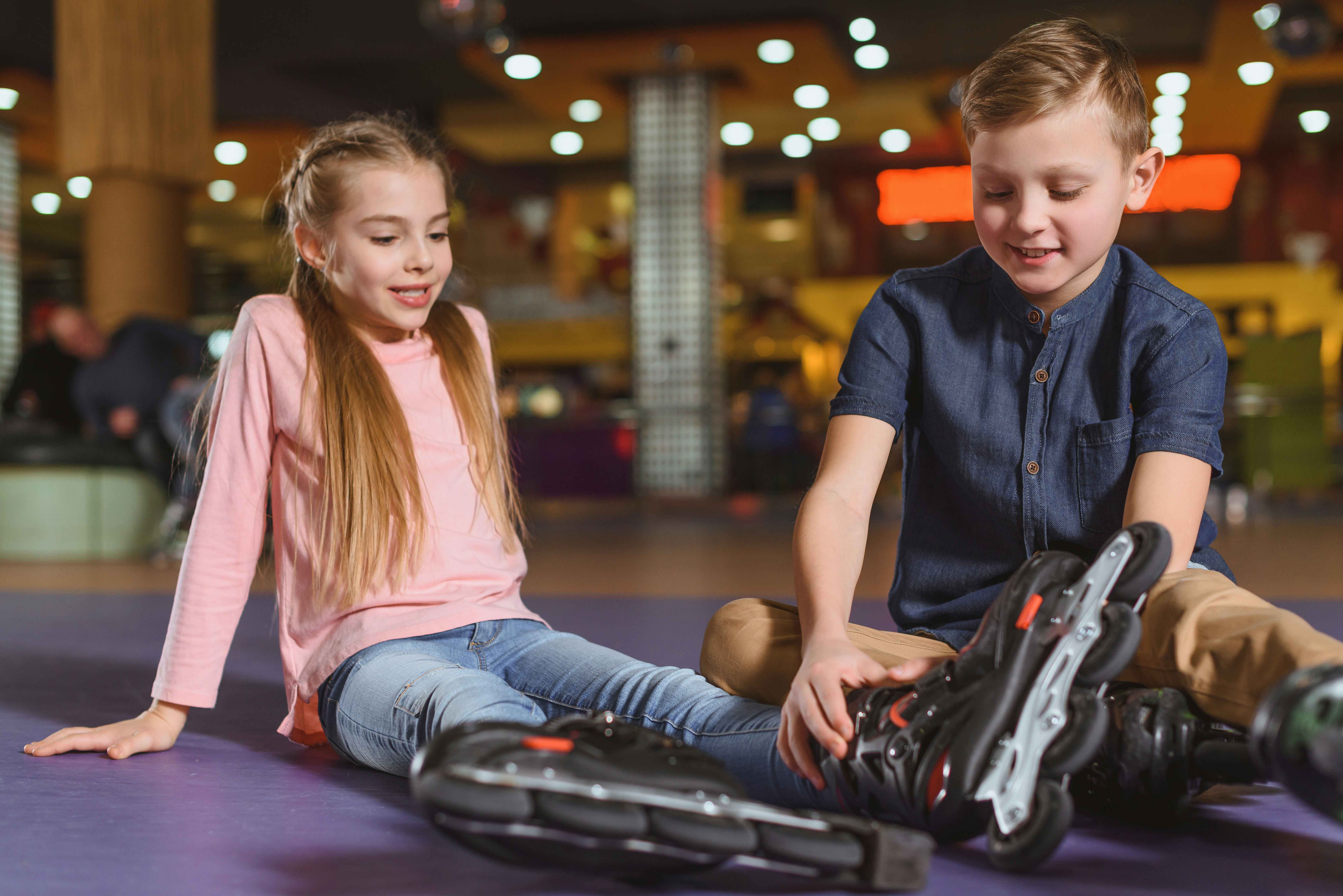 Two kids sitting on the floor of a roller rink with their rollerblades on.