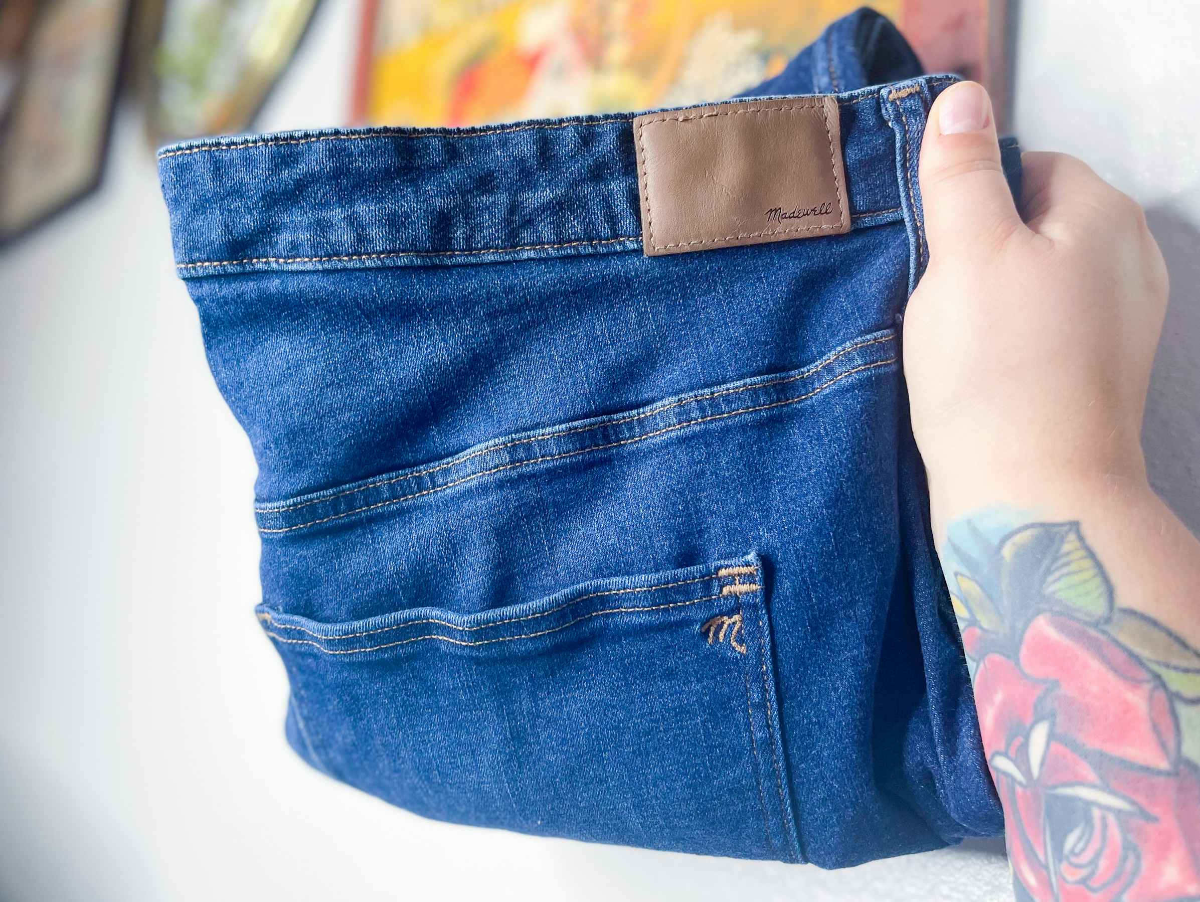 A person's hand holding up a pair of Madewell jeans in their home.