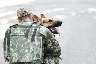 Member of the US Armed Forces dressed in a uniform kneeling down, hugging a German Shepherd dog.
