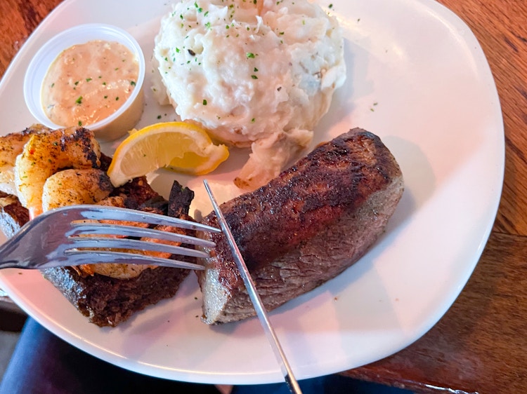 Someone using a knife and fork to cut into a steak on a plate with shrimp and mashed potatoes at Outback Steakhouse.