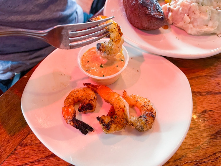 Someone using a fork to dip a shrimp into a dipping cup on a plate next to more shrimp at Outback Steakhouse.