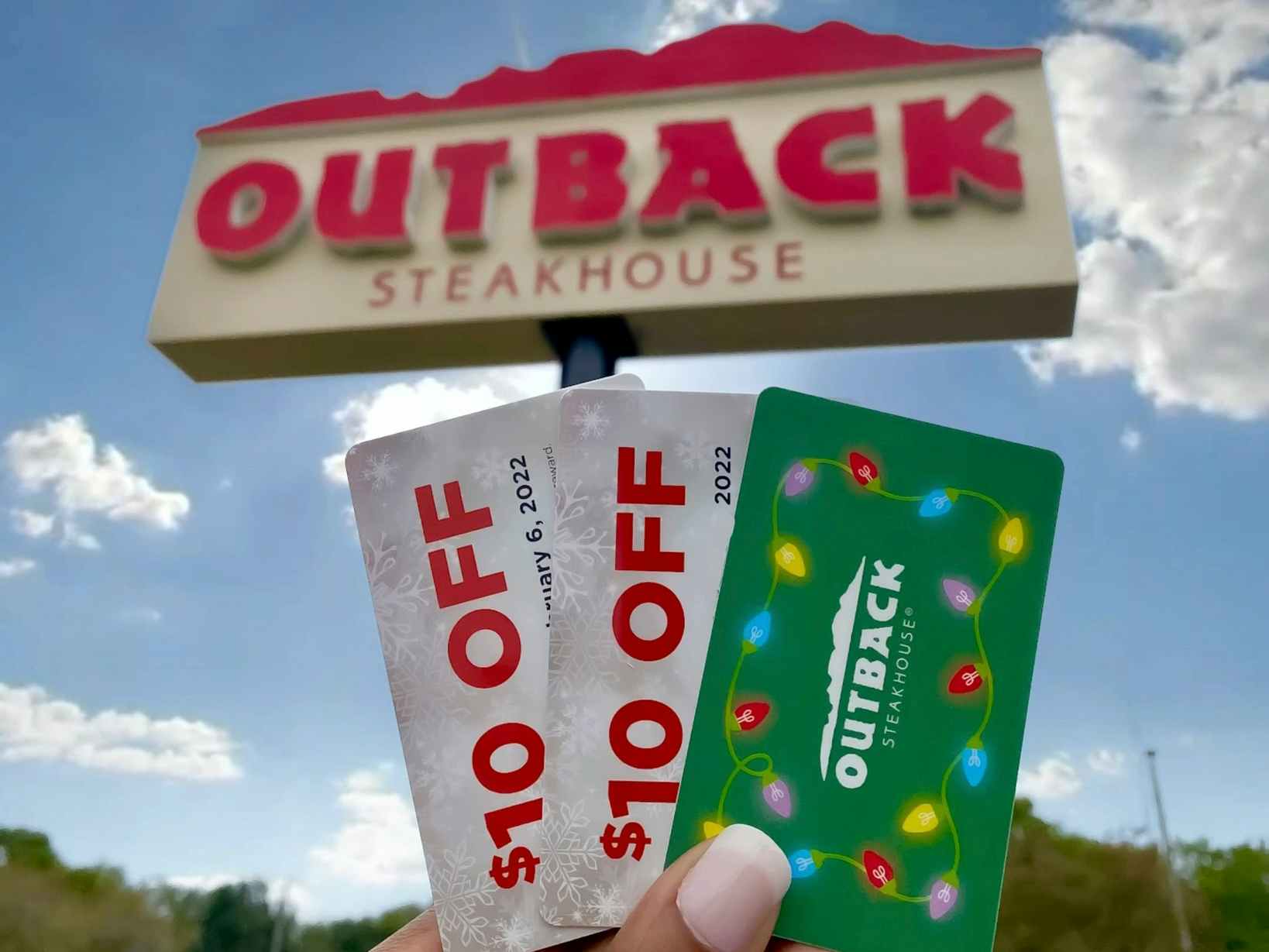 An Outback Steakhouse gift card and two bonus $10 OFF cards being held in front of an Outback Steakhouse sign.