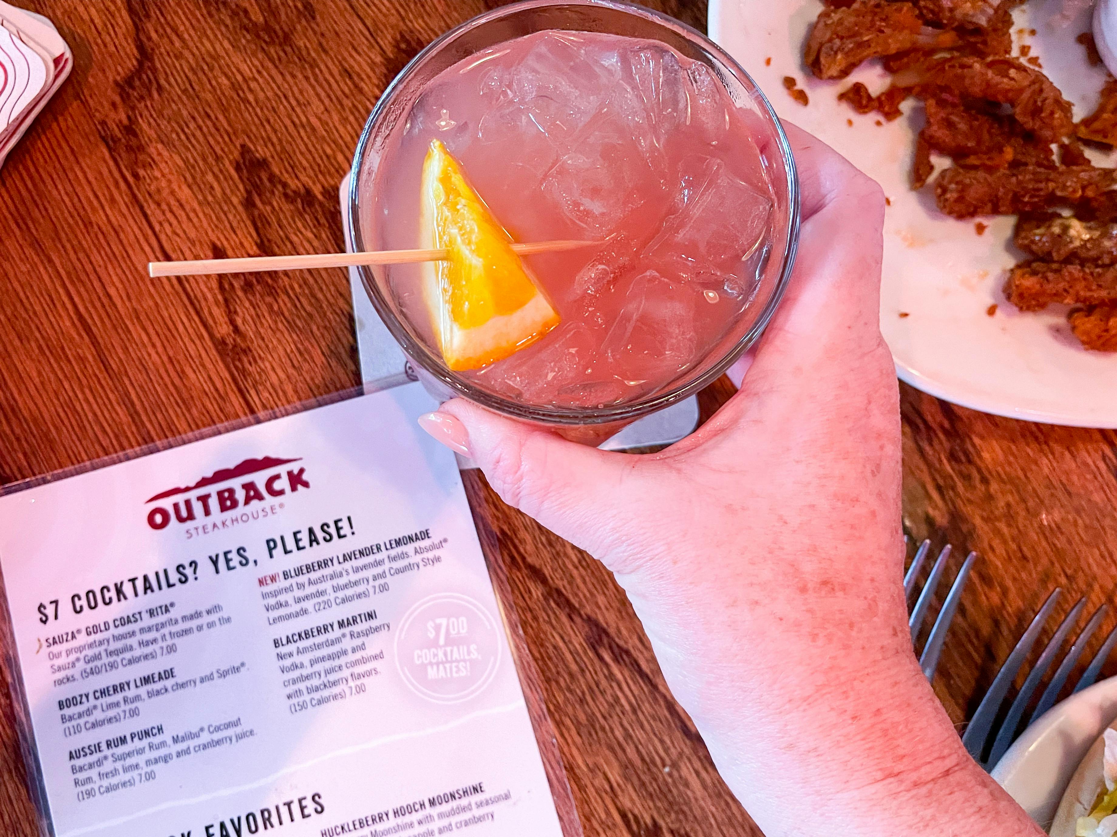A person's hand grabbing a cocktail drink sitting on a table next to an Outback Steakhouse drink menu.