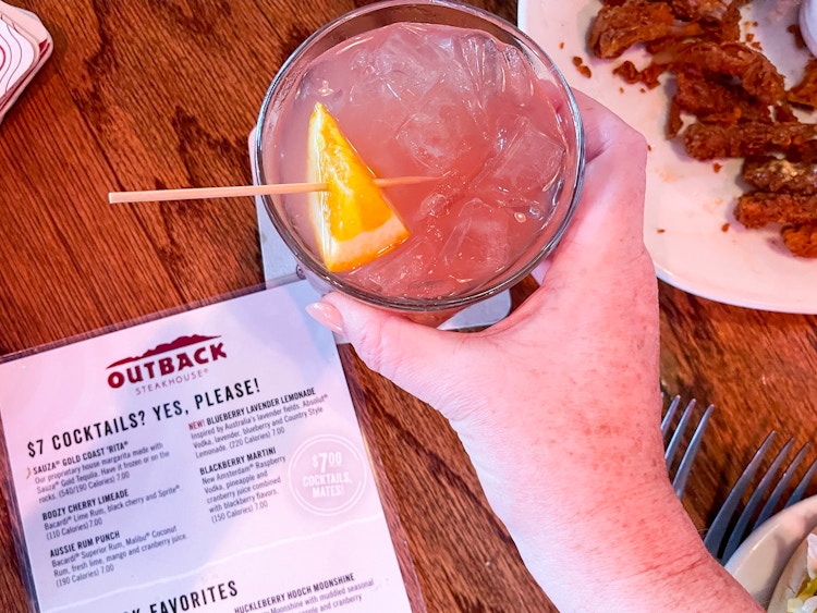 A person's hand grabbing a cocktail drink sitting on a table next to an Outback Steakhouse drink menu.