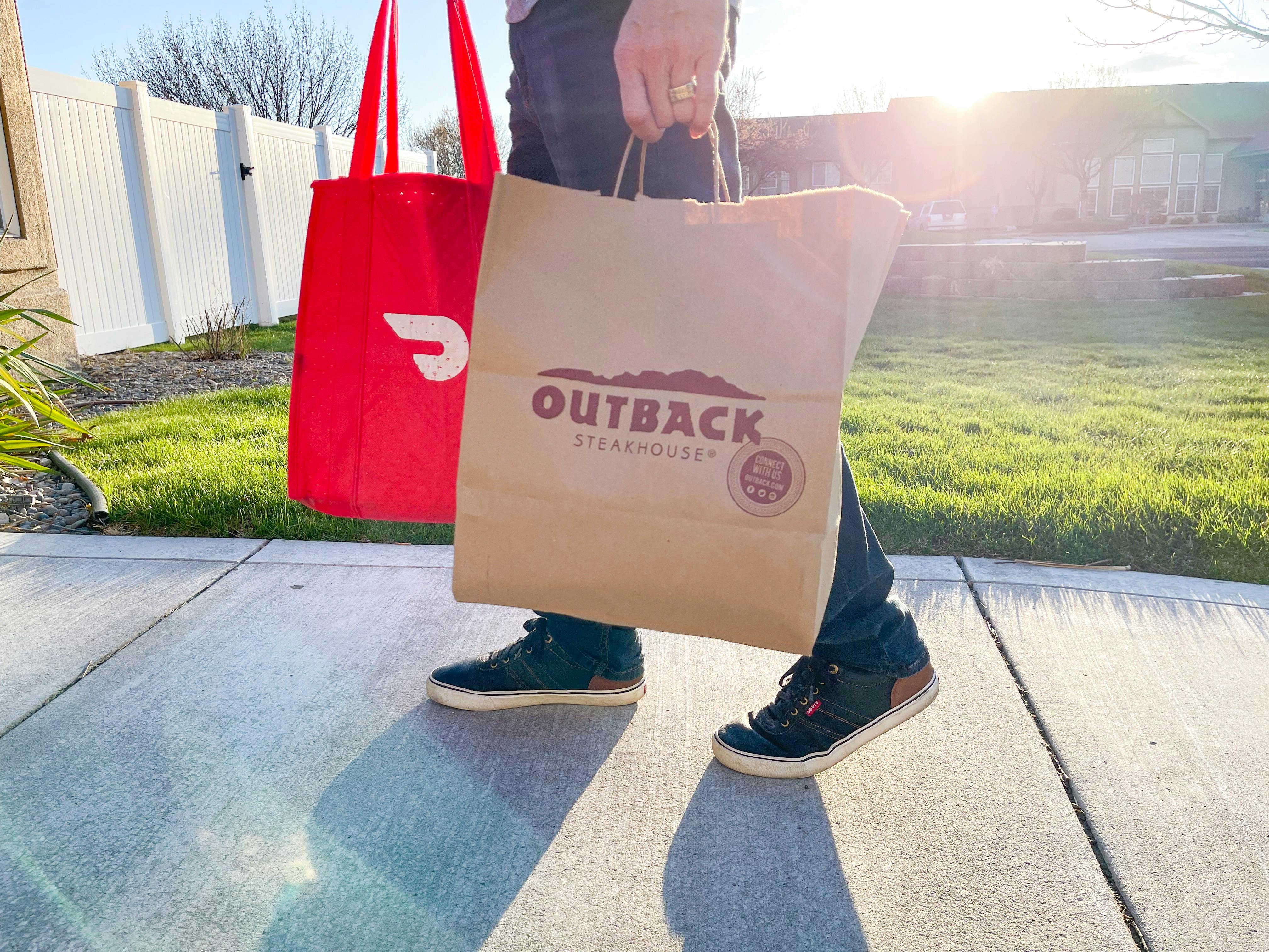 A person holding a DoorDash delivery bag and an Outback Steakhouse takeout bag walking down a sidewalk.