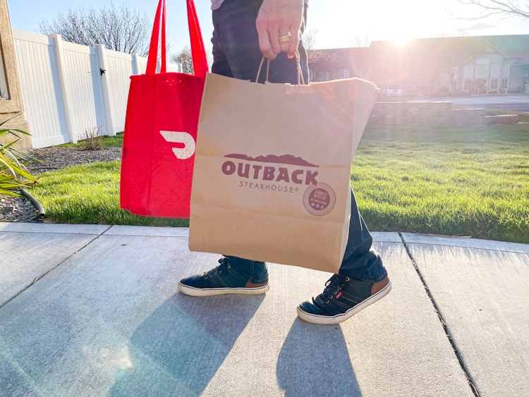 A person holding a DoorDash delivery bag and an Outback Steakhouse takeout bag walking down a sidewalk.