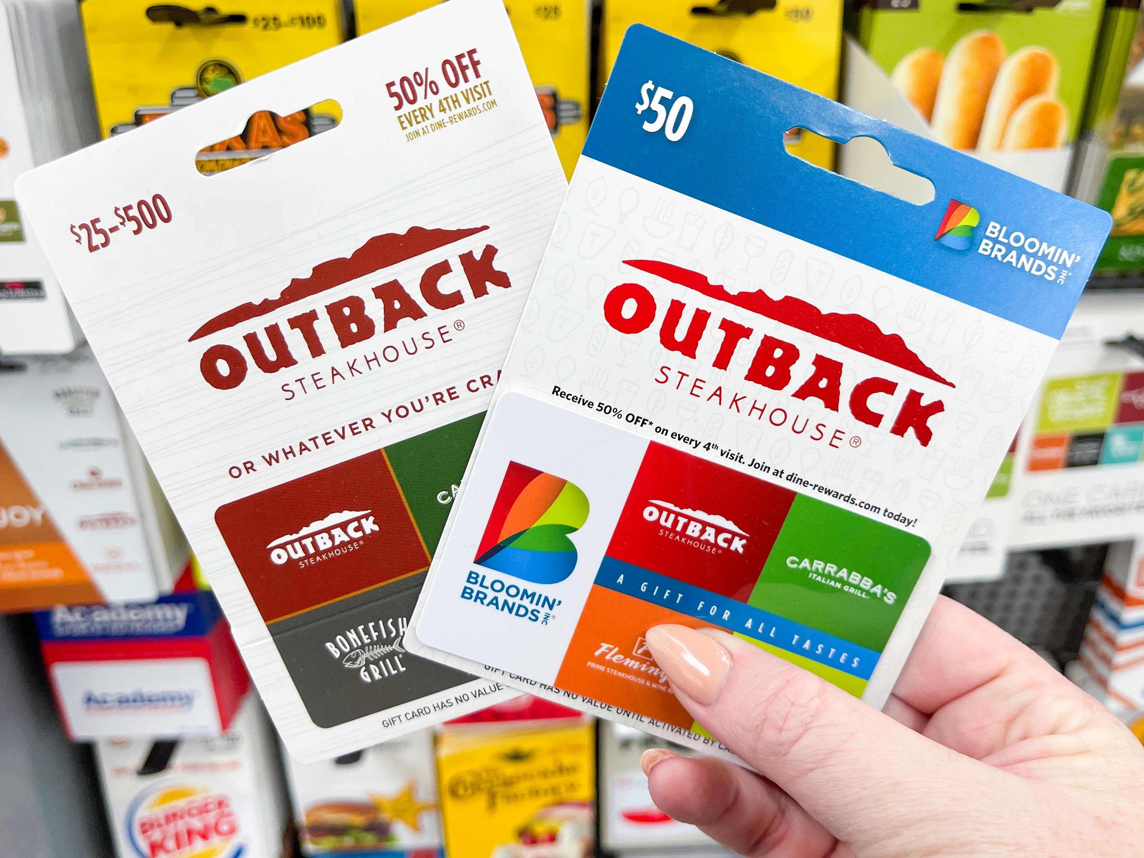 A person's hand holding two Outback Steakhouse gift cards in front of a display of gift cards.
