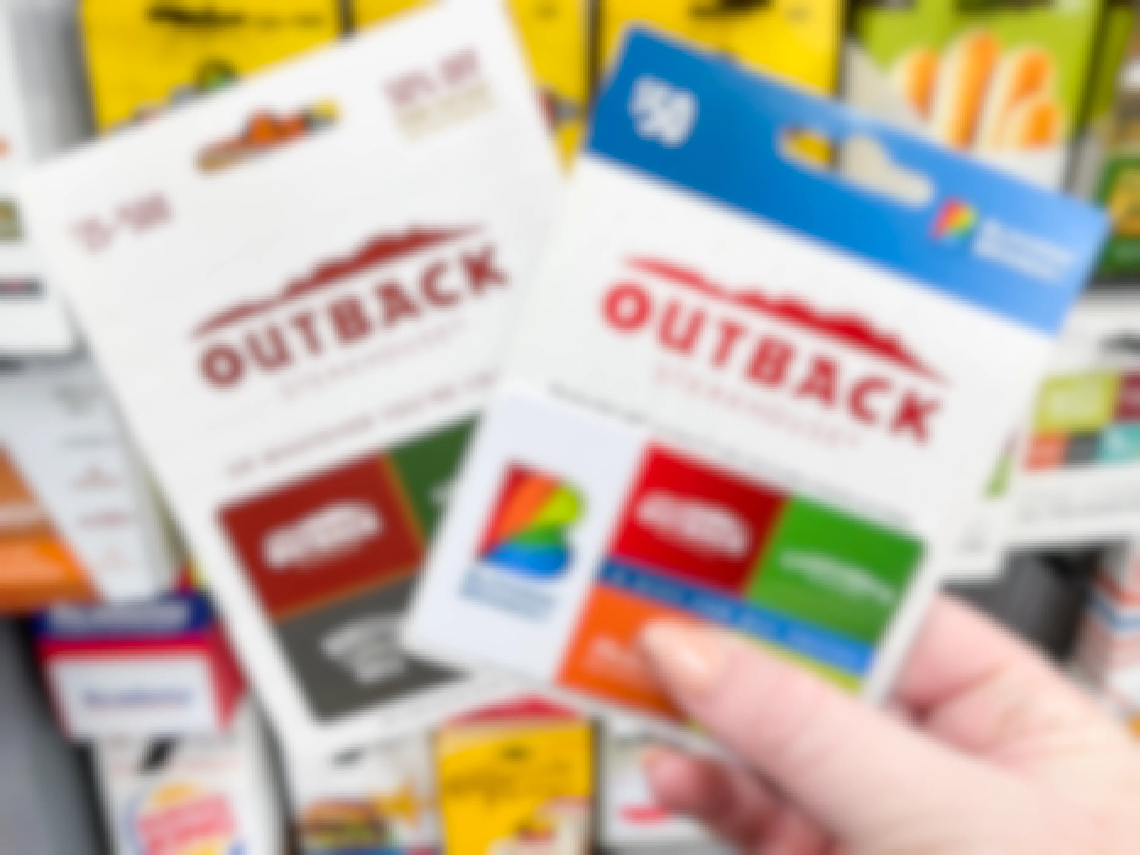 A person's hand holding two Outback Steakhouse gift cards in front of a display of gift cards.
