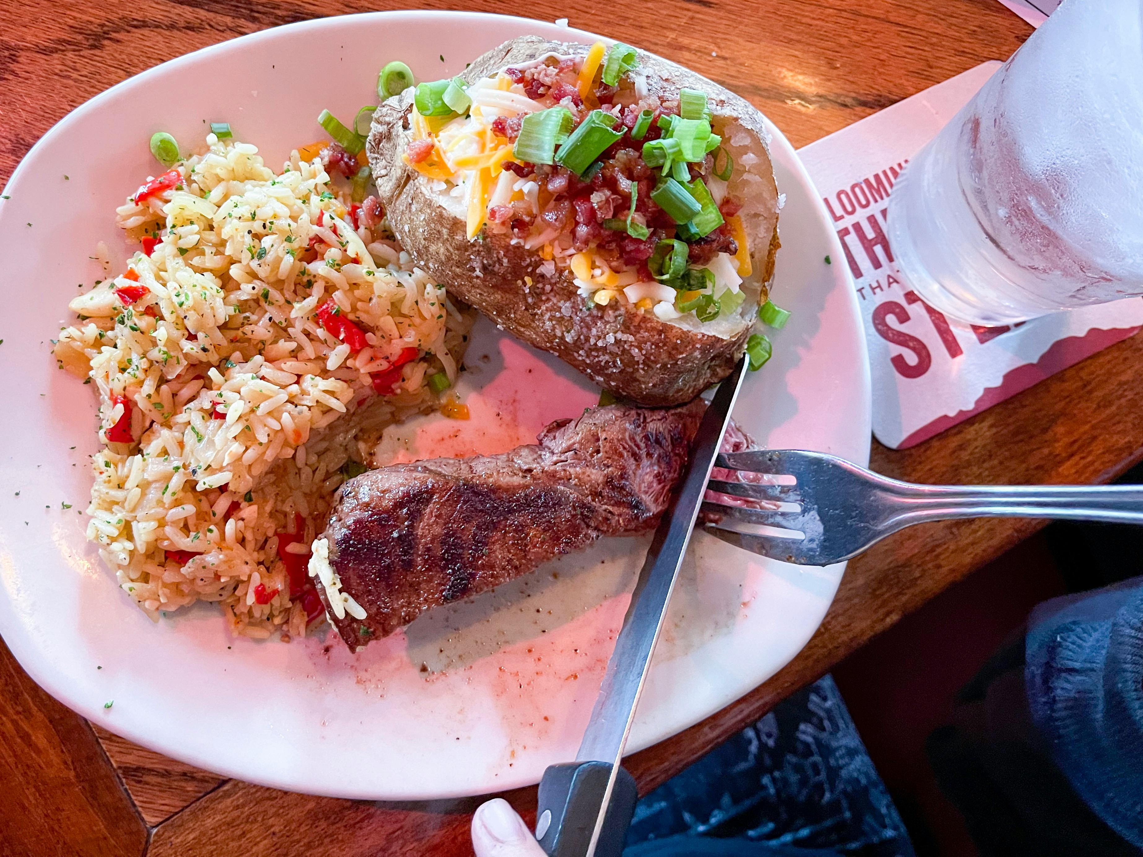 Someone using a fork and knife to cut into a steak on a plate with rice and a loaded baked potato at Outback Steakhouse.