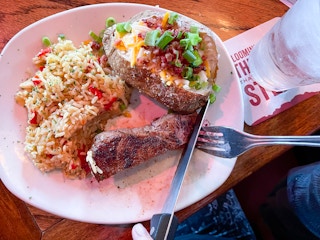 Someone using a fork and knife to cut into a steak on a plate with rice and a loaded baked potato at Outback Steakhouse.