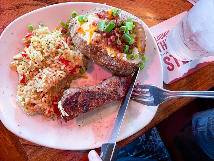 Someone using a fork and knife to cut into a steak on a plate with rice and a loaded baked potato at Outback Steakhouse.