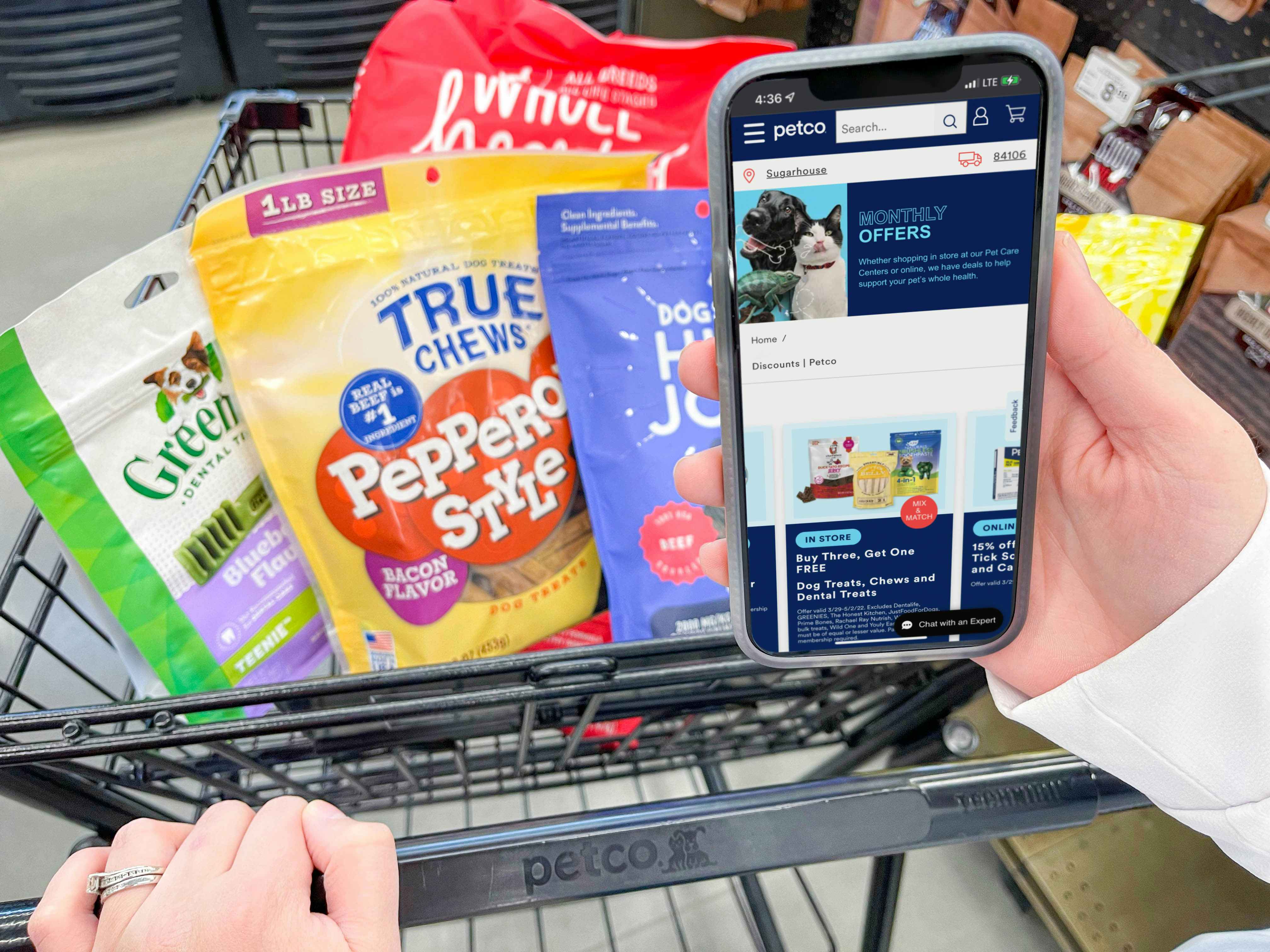 a person holding a cellphone with petco monthly coupons on screen while pushing cart with dog treats 