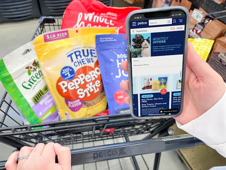 a person holding a cellphone with petco monthly coupons on screen while pushing cart with dog treats 